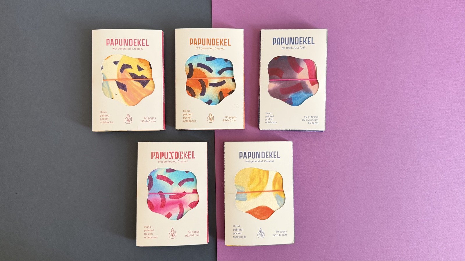 Five PAPUNDEKEL notebooks in their paper sleeves, each with a different hand-painted cover, on grey and purple paper