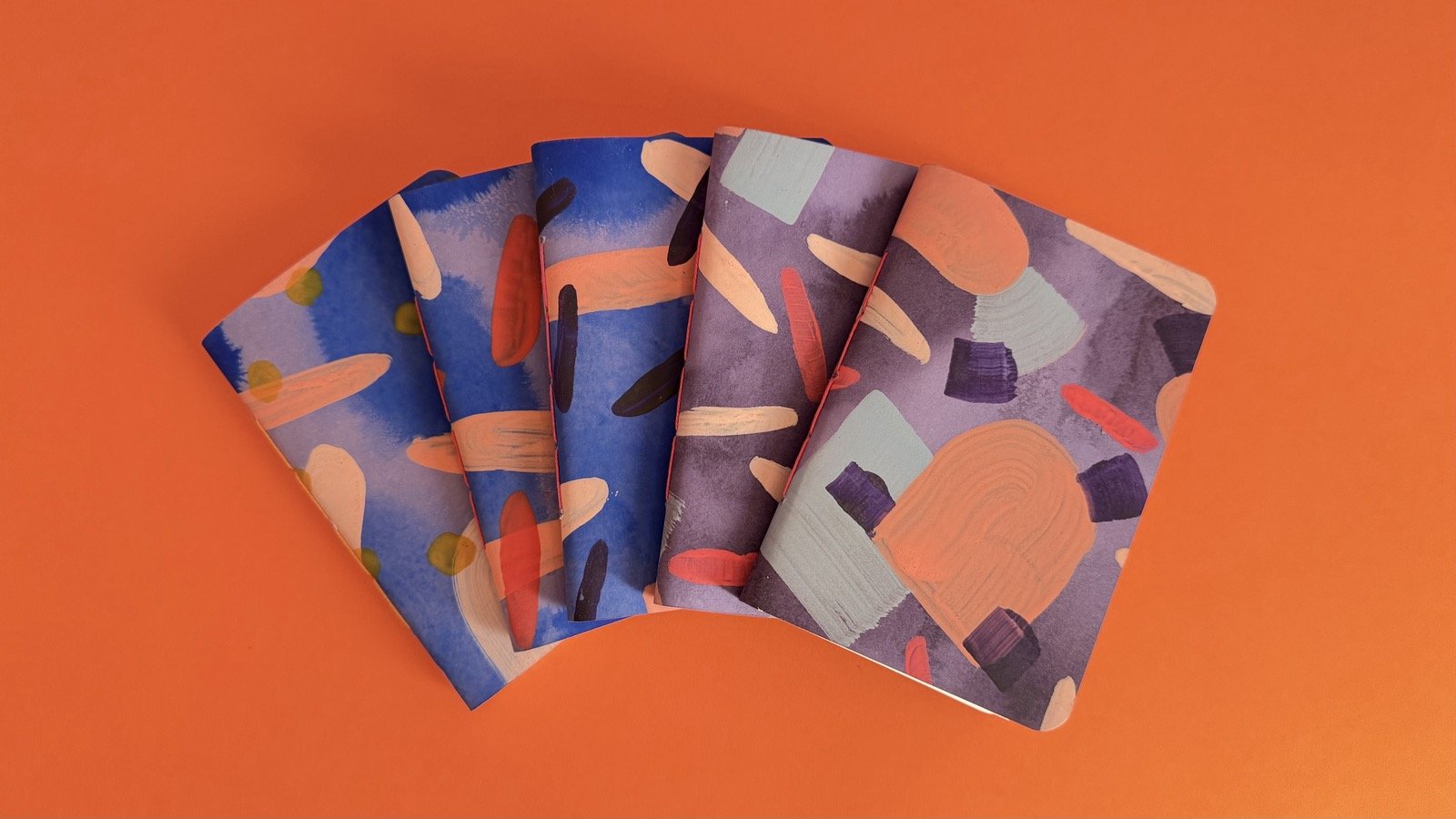 A fan of hand-painted notebooks in blue, purple and peach on an orange background