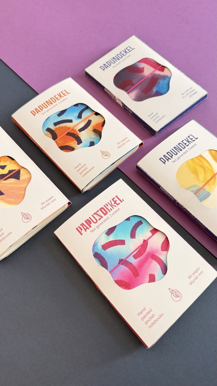 PAPUNDEKEL notebooks in their sleeves, scattered on purple and grey paper