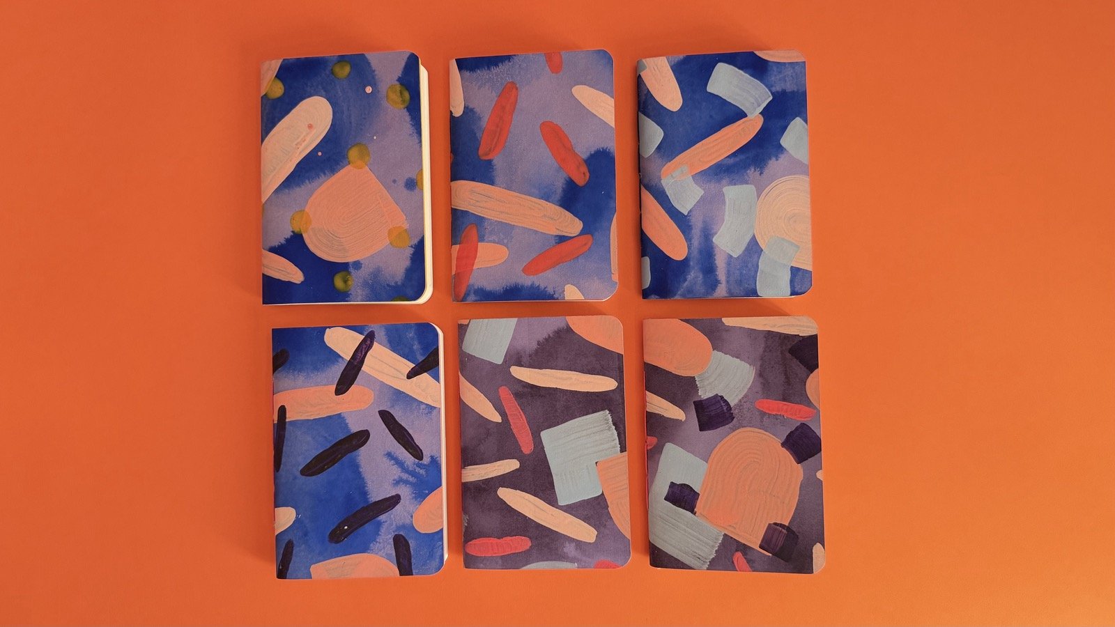 Six hand-painted notebooks laid out in two rows on an orange background